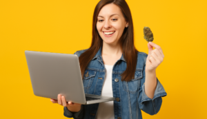 Smiling woman holding cannabis and laptop against a yellow background, representing excitement about buying BC pot online